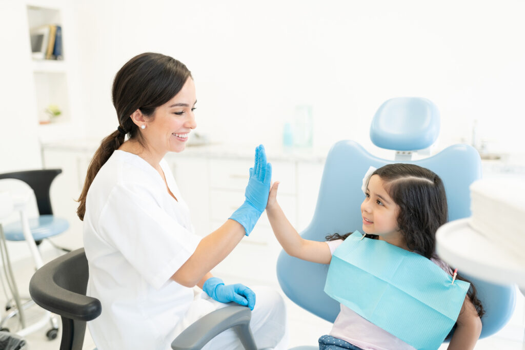 Cute Hispanic girl high-fiving with female dentist during checkup at dental clinic
