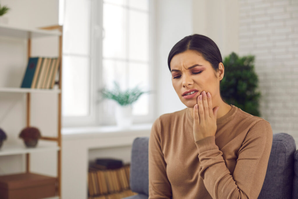 Woman suffering from toothache and touching cheek with closed eyes and grimace of pain. Young lady suffering from inflamed tooth nerve or teeth sensitivity. Concept of people having dental problems